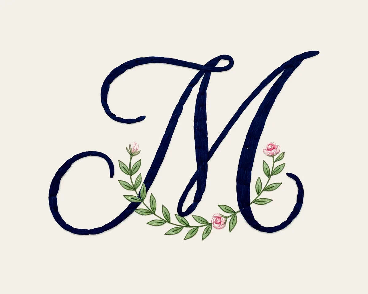 A navy script letter M with a small wreath of green leaves and pink rosebuds on cream paper